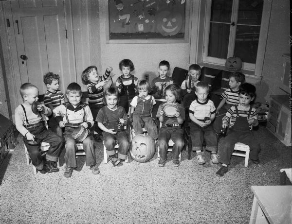 Group portrait of 13 preschool children, sitting and holding rhythm band instruments. There is a Halloween jack-o-lantern placed in the center of the group. The nursery school was in the Salvation Army building at 121 W. Wilson Street, and was a cooperative operated by the parents and administered by the Madison Board of Education.