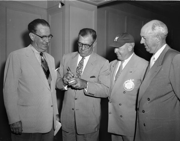 Charlie ("Jolly Cholly") Grimm, manager of the Milwaukee Braves, is shown second from left autographing a baseball before the Rotary luncheon at the Loraine Hotel in the Crystal Ballroom.  Surrounding Grimm, left to right, are Joseph (Roundy) Coughlin, <i>Wisconsin State Journal</i> columnist, Maj. Gen. Ralph Olson, Wisconsin adjutant general (wearing baseball cap), and Duane Bowman, president of the Wisconsin State League.