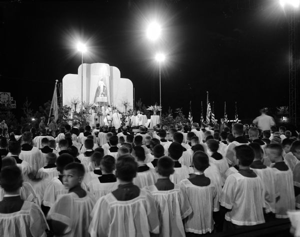 Over fifty altar boys kneel before a stage decorated with a statue of Mary, with flower arrangements and United States flags. Bishop William P. O'Connor of Madison and Bishop Raymond Hillinger of Rockford, Illinois preside.