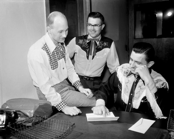 Three male square dance callers wearing western shirts sit around a desk.
