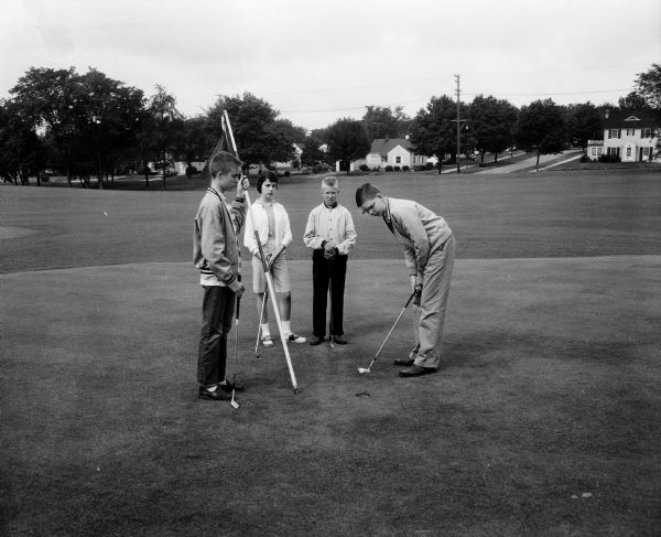 Golf Lessons for Children at Glenway Golf Course Four golfers take advantage of special rates offered to those who follow all the rules of golf. Don Gilpin (right) is putting while Bobby Bolger (left) is holding out the flag. Pat Mitchell (female) and John Dyreson are looking on. Three houses are in the background.