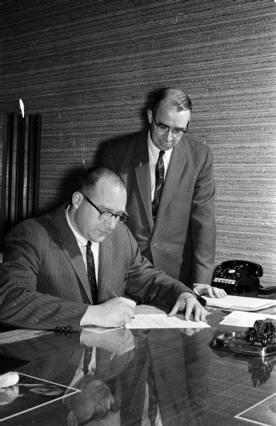 Two incorporators of the Madison Junior Achievement Corporation sign the papers that will put teen-age boys and girls into their own profit-making businesses. Seated is John T. Lunenschloss, president of Madison-Kipp Corporation. Standing is William McNamara, a director of Madison Gas and Electric Company.