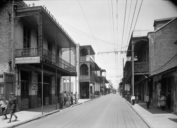 New Orleans street scene. Pedestrians can be seen on the sidewalks, and a streetcar can be seen in the distance. "Moskau Cabinet Works, door and window screens" can be read on building on the left side of the street.