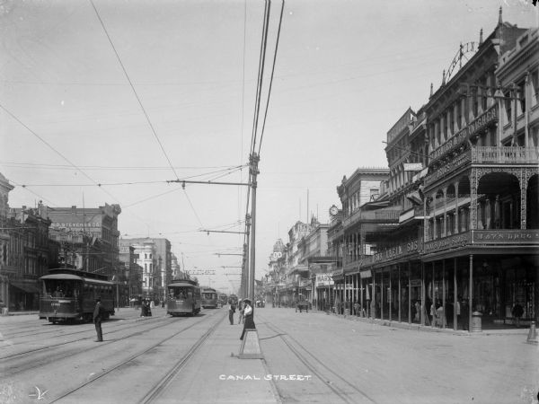 View down Canal Street in New Orleans. View is toward the lake from Camp/Chartres Streets. The first building on the right reads "May's Drug Store". The two closest streetcars are both "Palace" cars. The car to the left is "Palace" car 036, on the riverbound inner track. The other "Palace" car is on the lakebound inner track. Caption reads: "Canal Street."