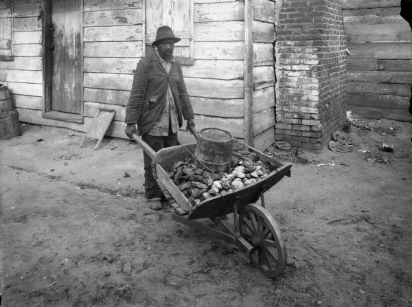 An African American vendor of oysters and clams is shown pushing his wheelbarrow. A building with an exterior brick chimney is behind him.