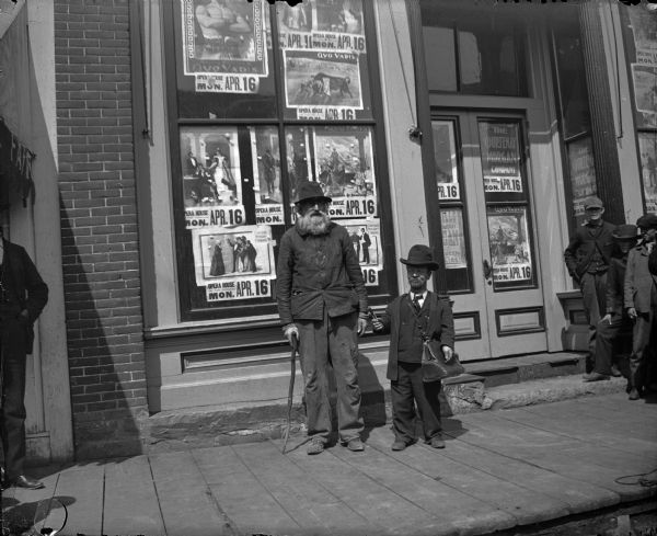 Two men posing standing in front of the Fair Store on Main Street. The man on the right is a dwarf and he is holding a cigar. Posters in the windows advertise "Quo Vadis at the Opera House on Monday, April 16" by the Courtenay Morgan Company.