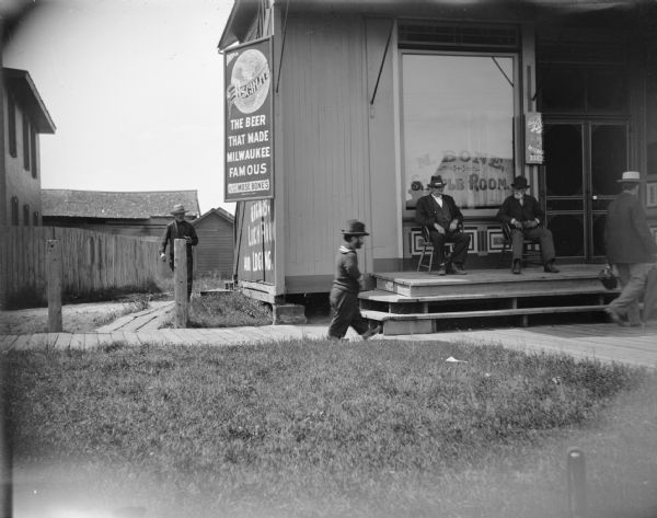 Men at the Mose Bone Sample Room. Two men are sitting on chairs on the porch in front. A male dwarf is walking on the board sidewalk in front of the saloon, which is north of the railroad depot. A sign on the building advertises "Drink Schlitz, the Beer that Made Milwaukee Famous, For Sale at Mose Bones." There is a sign painted on the side of the building advertising "Cigars, Lunch Room, and Lodging."