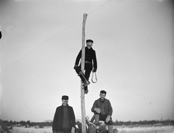 Man posing on a telephone pole with climbing equipment, with two men standing below.