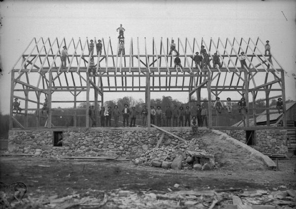 Barn raising. The foundation is constructed from local fieldstone or "pudding stone," found in the nearby fields. A large group of men are is posing holding an infant, and a young boy is standing and holding the hand of another man.