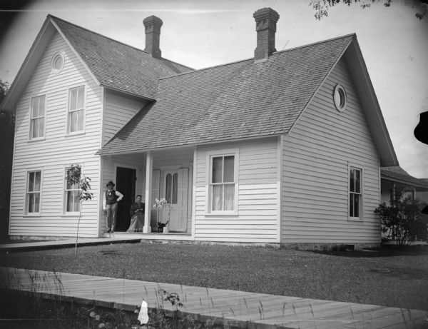 Man, woman, and cat on the porch of a frame home, with board sidewalk in foreground.  This is probably Mr. and Mrs. Bartell Giroux. He was a tinsmith.
