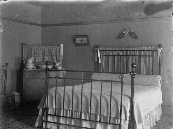 View of a metal post double bedstead against a wall, and short dresser in the corner, and a chair on the far left beside a chamber pot. A water pitcher, lavatory bowl, and two other vessels are sitting atop the dresser. A framed picture is hanging from the picture rail to left of bedstead, and two crossed fans are hanging over the bed. A portion of stovepipe is on the right. A patterned rug or carpet is on the floor.
