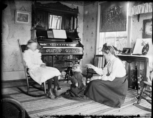A girl sits in a rocking chair as a young woman commands a dog who is dressed in dark clothing to sit. A piano or organ is in the background.