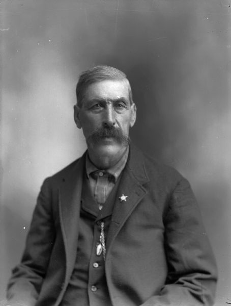 Studio portrait of a seated man with a moustache. He is wearing a suit jacket with a star-shaped pin on the lapel, and a watch fob on his vest.