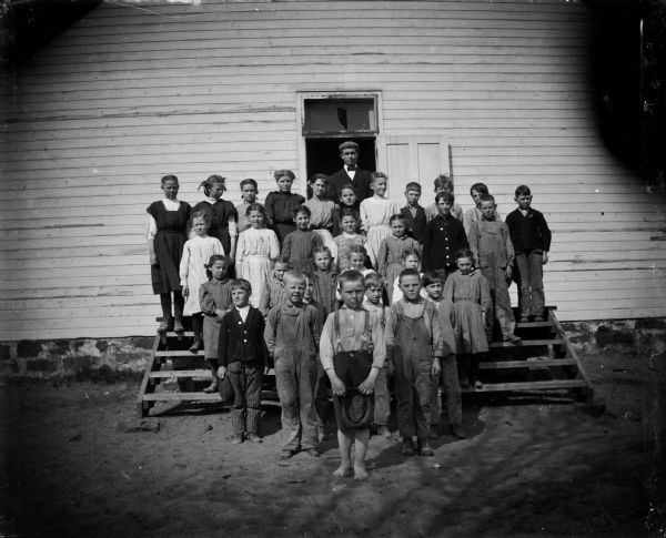 Group of children and a man, probably a teacher, posed standing on the steps of a building, probably a schoolhouse.	