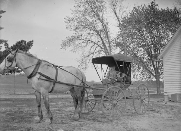 Woman and Girl in Carriage A woman and young girl sit in a carriage that is pulled by a horse. The corner of a building is on the right.