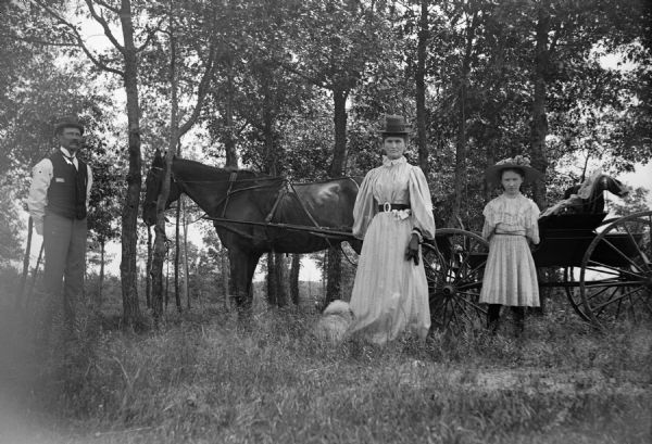 Man, Woman and Girl Posed in front of Carriage A man, woman and girl stand in front of a carriage pulled by a horse in the woods.