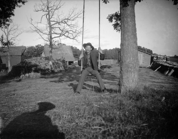 A man, possbly Andrew Larson (?) of Hoffman Creek, sits on a tree swing. The shadow of the photographer is visible on the grass.