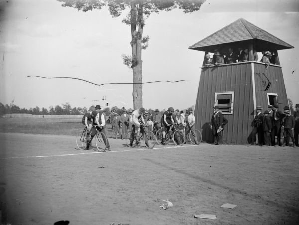 Four cyclists await the start of a race at the Jackson County Fairgrounds. There is a viewing pavilion on the right.