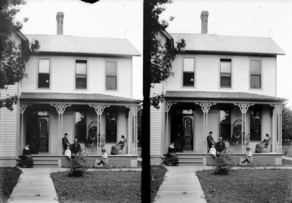 A stereograph from front walk of a family posing on a porch. A young child is riding a tricycle on a board sidewalk in front of the porch.