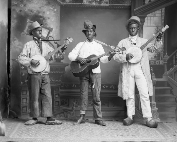 Three musicians, two males with banjos and possibly a woman with a guitar, pose in blackface and costumes for a studio portrait in front of a painted backdrop.