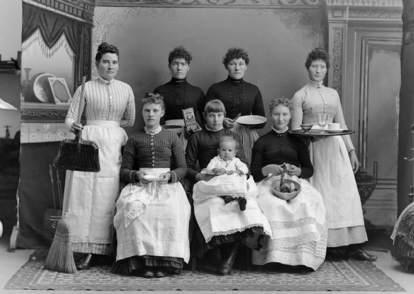The household staff (probably Swedish) for one family home, including laundresses, cooks, parlor maids, and scullery girls with their various work utensils. One woman is holding an infant. They are posing in front of a painted backdrop.