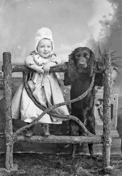 Portrait of a Small Girl and Dog A young girl stands on a wooden fence in front of a painted backdrop. A dog, possibly a Spaniel, stands on its haunches and rests its front paws on the fence.