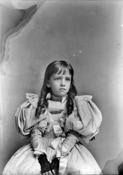 Girl in Puffy Sleeved Dress A studio portrait of a young girl with curled dark hair, sitting on a chair in a puffy-sleeved dress with a bow around her waist. She is wearing fingerless gloves.