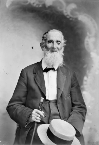 Studio Portrait of Elderly Man A studio portrait of a seated elderly man with a long beard posing in front of a painted backdrop,holding a cane and hat.