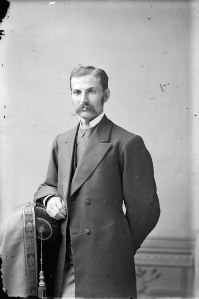 Studio portrait of a standing man with his arm on a stuffed chair, possibly a self-portrait of Van Schaick. He has a moustache, and is wearing a long jacket over a vest and high-collared shirt.