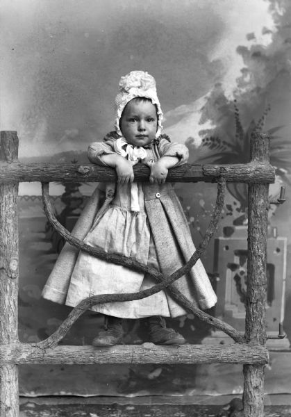 Portrait of Young Girl on Fence Studio portrait of a young girl wearing a bonnet, and a long coat over a dress, perched on a wooden fence in front of a painted backdrop.
