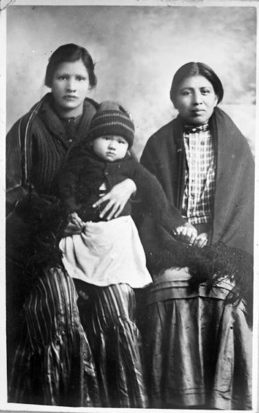 Two Young Ho-Chunk Women with Child Copy photograph of a studio portrait of two young Ho-Chunk women sitting and wearing wool shawls over long skirts or dresses. Names (l to r) Maude Looking Glass-Browneagle, Decorah, her daughter, Elsie Decorah, and Annie Massey (daughter of William Massey).