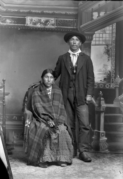 Studio portrait in front of a painted backdrop of a couple, who are the parents of Tom (?) Wallace. The woman is sitting with a blanket around her shoulders, and is wearing beaded necklaces and ear bobs. Her husband is standing next to her wearing a hat, suit jacket, vest, and trousers. Amos Wallace (Potawatomi or Menominee) and his wife, Annie Bearchief, also the mother of Joe Green.