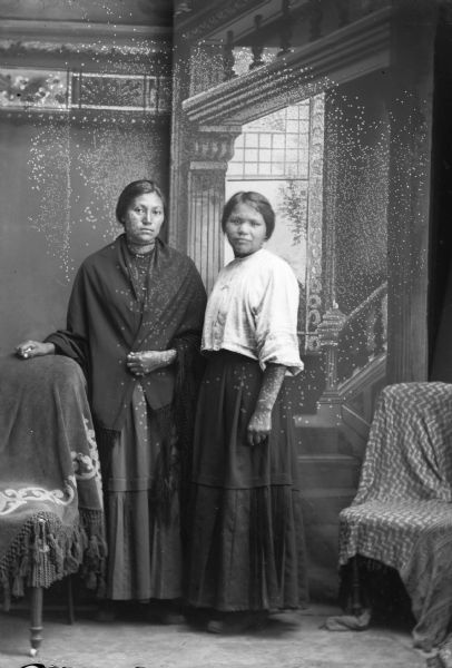 Portrait of Lucy Bear and Connie Blackhawk Studio portrait of two Ho-Chunk women posing standing in front of a painted backdrop. They are wearing contemporary dresses and short hair. The woman on the left, identified as Lucy Bear, the wife of George Bear, wears a shawl over her shoulders, and Connie Blackhawk on the right has a light-colored blouse.