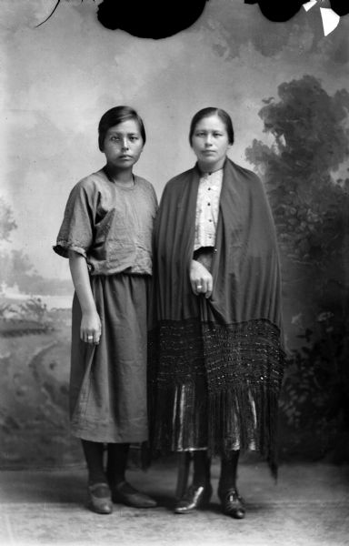 Studio Portrait of Alice Red Bird and Marie Johnson Studio portrait of two young Ho-Chunk women posing standing and wearing contemporary dress in front of a painted backdrop. The woman on the right is wearing an eight row jet bead silk shawl. Identified as Alice Red Bird on the left, and Marie Johnson on the right.