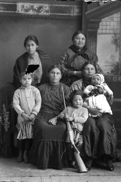 Portrait of Four Ho-Chunk Women and Three Ho-Chunk Children Studio portrait in front of a painted backdrop of two Ho-Chunk women posing sitting, one holding a small child, two Ho-Chunk children standing, one holding a rifle, in front of two Ho-Chunk women standing behind the group. All are wearing contemporary clothing. The woman standing on the left is Nellie Winnesheik Red Cloud, the woman standing on the right is the wife of St. Cyr, and the woman sitting on the left is Mary Dixon.