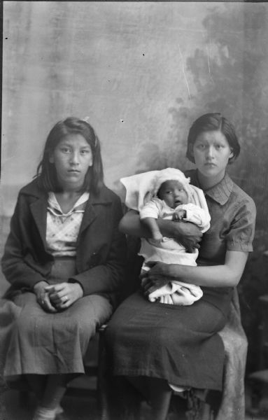 Studio Portrait of Frieda and Stella Stacy with Infant Studio portrait of two young Ho-Chunk women sitting in front of a painted backdrop. They are wearing contemporary dress. The woman on the right is also holding an infant. Identified as Frieda Stacy on the left and Stella Stacy on the right.