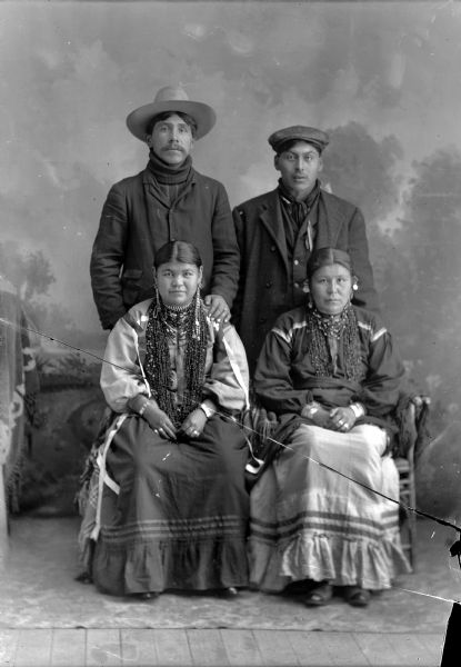 Studio Portrait of Two Ho-Chunk Couples Studio portrait in front of a painted backdrop of two Ho-Chunk women posing sitting and wearing several necklaces and earrings, in front of two Ho-Chunk men wearing suits and hats. Names, standing (l to r): Amos Wallace (Wee Kah), George Otter (Hay Cho Kah). Sitting (l to r): Belle Youngswan, Standingwater, Whitepigeon, Wallace (Wa Nek Monk E Kay Win Kah) and Annie Youngswan, Littlesnake, Otter (Wau Kon Chaw Mon Nee Win Kah).