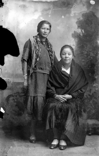 Studio Portrait of Two Ho-Chunk Women Studio portrait in front of a painted backdrop of a Ho-Chunk woman posing standing, with a scarf around her shoulders, and another Ho-Chunk woman posing sitting, wrapped in a dark-colored fringed shawl.