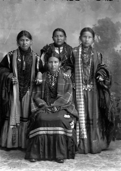 Studio portrait in front of a painted backdrop of a Ho-Chunk woman posing sitting in front of three Ho-Chunk women posing standing, all wearing several necklaces, earrings, and rings. The women in the center have shawls around their shoulders, and the women on the left and right have their hands on the seated woman's shoulders, and shawls over their outer arms.
