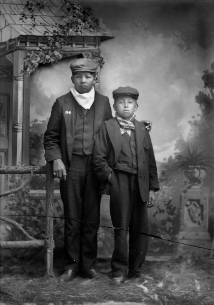 Studio Portrait of Two Ho-Chunk Boys Studio portrait in front of a painted backdrop of two Ho-Chunk boys posing standing next to a prop wooden fence. They are both wearing suits, bandanas, and caps. The older boy on the left has his left hand on the younger boy's left shoulder, who has his right hand in his pocket.
