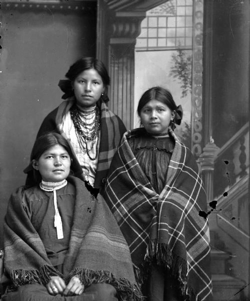 Portrait of Ethel Windblowe, Sarah Windblow, and Elizabeth Thunder Cloud Studio portrait of a Ho-Chunk woman posing sitting on the left, and two women posing standing in the center and on the right. They are in front of a painted backdrop, and are wrapped in shawls with ribbons in their hair. The women on the left and right are wearing several short necklaces, and the woman in the center is wearing several long necklaces. They are identified from left to right as: Ethel Windblowe sitting on the left, Sarah Windblowe standing center, and Elizabeth Thunder Cloud standing on the right.