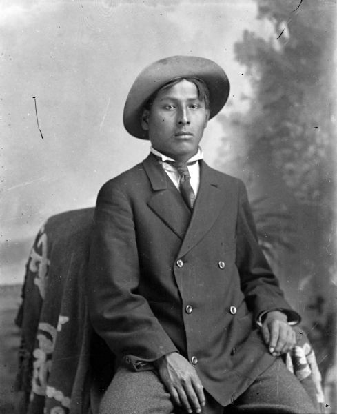 Portrait of a Young Ho-Chunk Man Posed Sitting Studio portrait of a young Ho-Chunk man posing sitting in front of a studio backdrop. He is wearing a double-breasted suit coat, necktie, and hat.