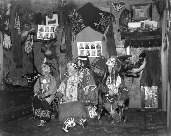 A European American woman and two boys posing sitting and a European American girl posing standing, all dressed in Ho-Chunk regalia. They are in a room decorated with Ho-Chunk artifacts and photographs, and the artifacts are probably from the collection of Tom Roddy.