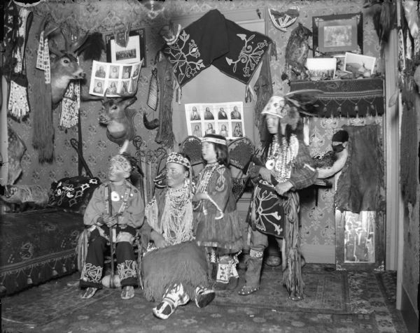 European American woman and boy posing sitting, and a European American girl and boy posing standing, all dressed in Ho-Chunk regalia, in a room decorated with Ho-Chunk artifacts and photographs. The artifacts are probably from the collection of Tom Roddy.