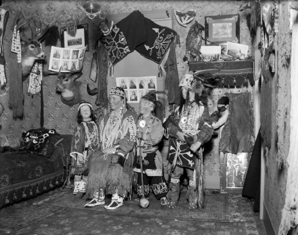 European American woman posing sitting, and a European American girl and two boys posing standing, all dressed in Ho-Chunk regalia. They are in a room decorated with Ho-Chunk artifacts and photographs. The artifacts are probably from the collection of Tom Roddy.
