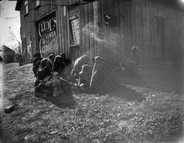Ho-Chunk women and children wrapped in shawls and sitting on the ground outside the livery.