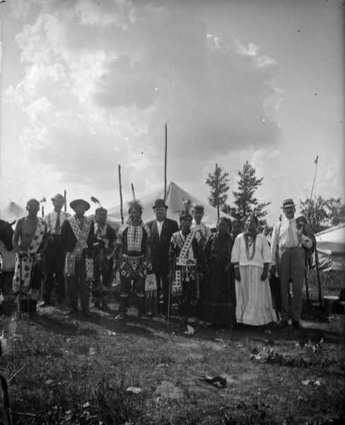 Several Ho-Chunk men, women, and a few European American men, posing standing in front of tents in a field and wearing regalia. They are members of the Blue Wing Band at Valley Junction on the Wyatt and Purdy Meadow.