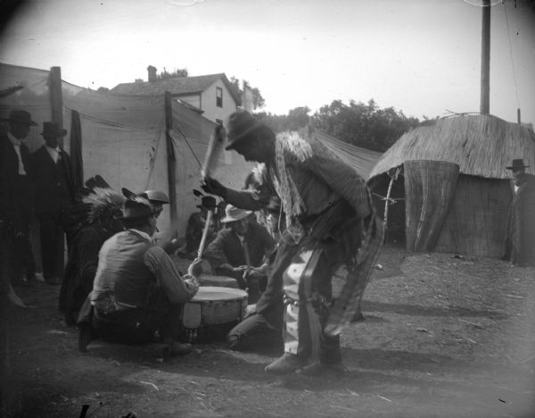 A Ho-Chunk man dressed in a mix of native and contemporary clothing dancing around singers seated around a drum. Ho-Chunk men are sitting and standing around a medicine drum, possibly the Dream Drum, in front of a cattail mat lodge in an area partitioned off by canvas barriers in front of a lodge. Identified as the Homecoming Powwow at the intersection of Main and Second Streets.