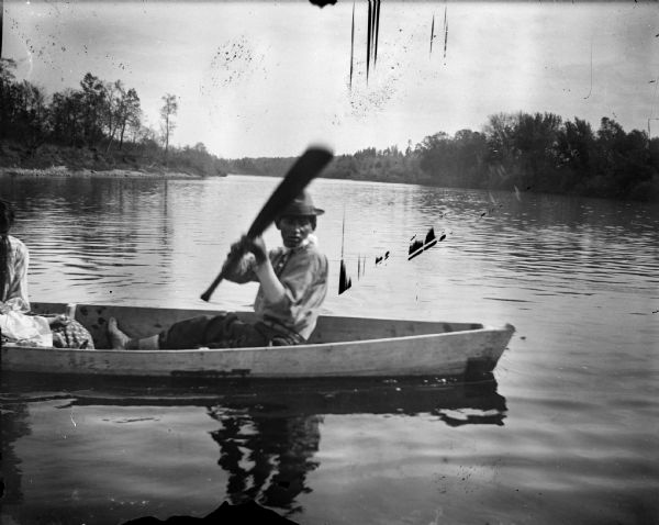 Ho-Chunk man in a canoe on a river holding up an oar. Just out of frame on the left is a woman, who is perhaps holding a child.