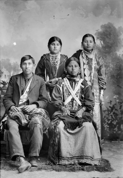 Studio Portrait of a Ho-Chunk Family Studio Portrait of a Ho-Chunk man, woman, and two girls in front of a painted backdrop. The Ho-Chunk man is posing sitting on the left and is wearing a bandoleer underneath a suit coat and has a shawl on his lap. The Ho-Chunk woman is sitting next to him on the right and is wearing many necklaces, earrings, rings, and bracelets and also has a shawl on her lap. The two girls standing behind the man and woman and are both wearing necklaces, earrings, bracelets, and shawls.